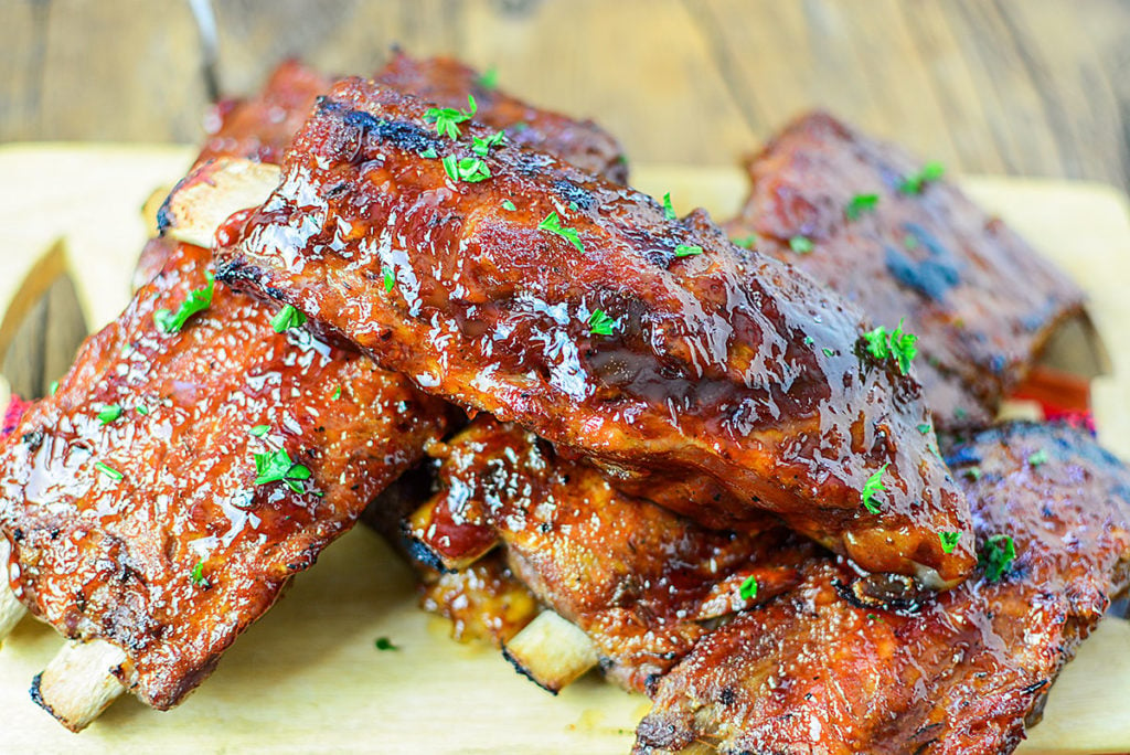 Close up image of 5 pieces of OVEN ROASTED LOW AND SLOW BBQ RIBS sitting on a wooden chopping board, garnished with some chopped parsley.