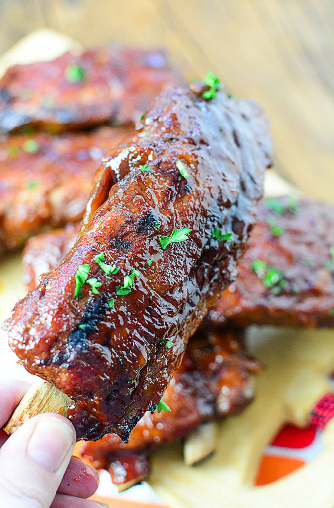 Close up image on a piece of OVEN ROASTED LOW AND SLOW BBQ RIBS being held by hand (where the thumb and other three fingers are index finger of a person is showing) At the background are 4 pieces of the BBQ ribs sitting on wooden chopping board and garnished with some chopped parsley.