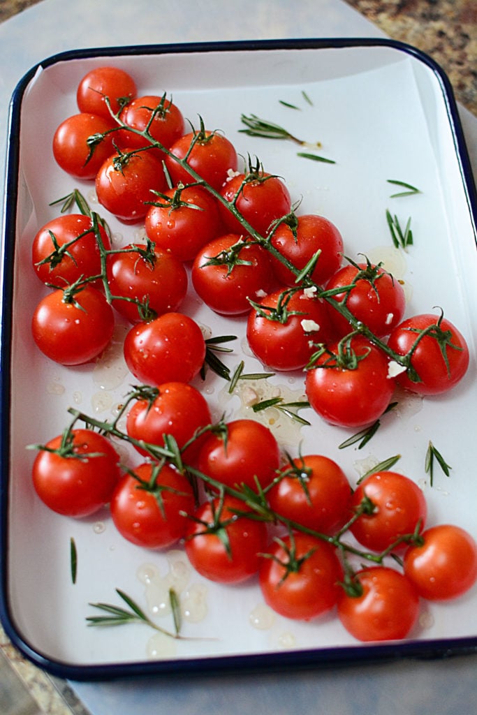 Fresh red tomatoes on a vine sitting in a white baking dish with oil and seasonings around it