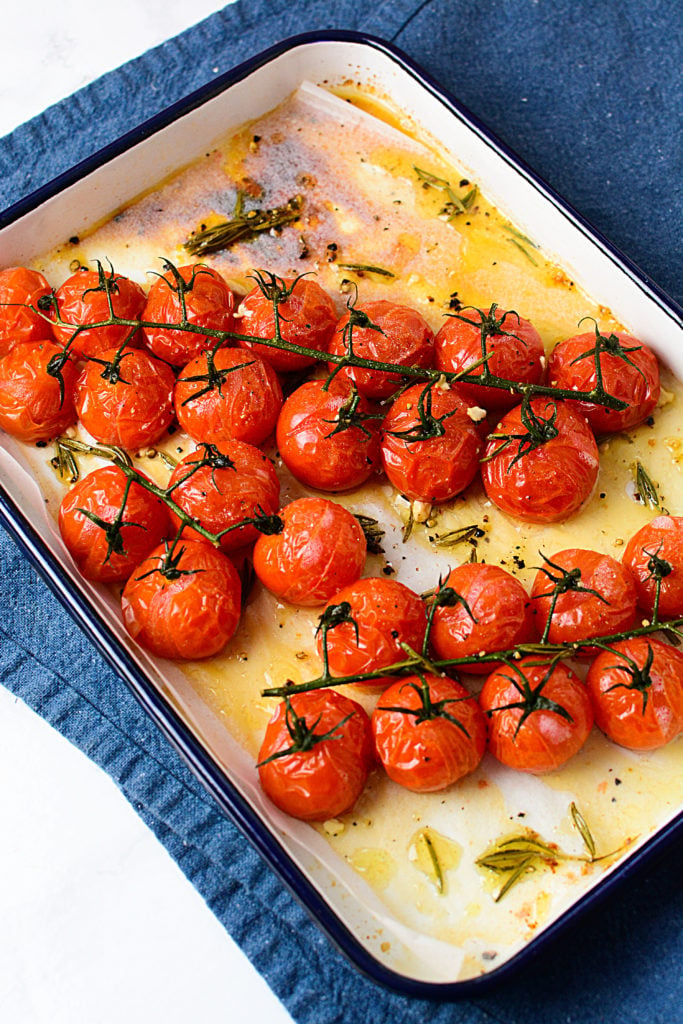 Roasted red tomatoes on a vine sitting in a white baking dish with oil and seasonings around it