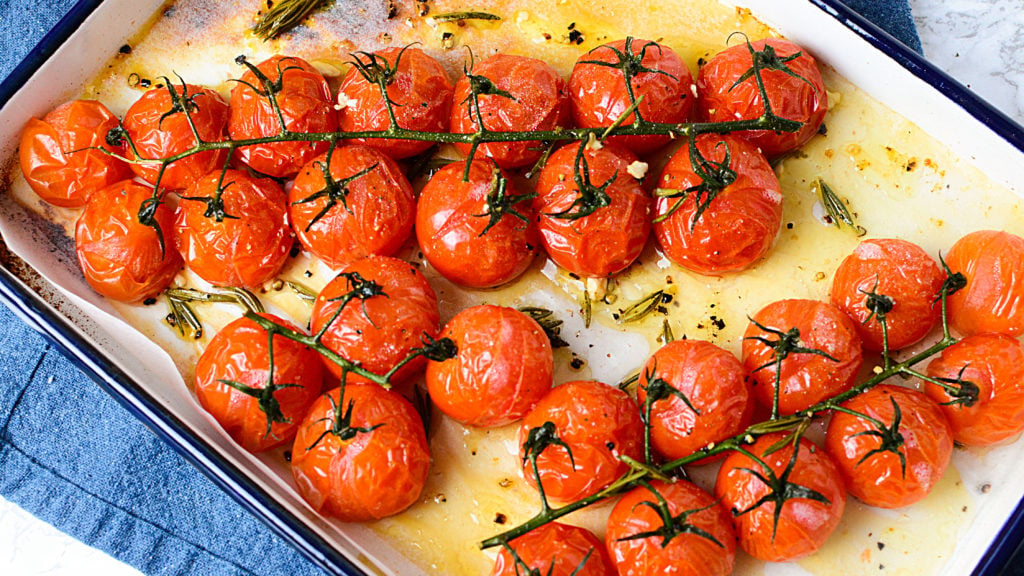 Roasted red tomatoes on a vine sitting in a white baking dish with oil and seasonings around it