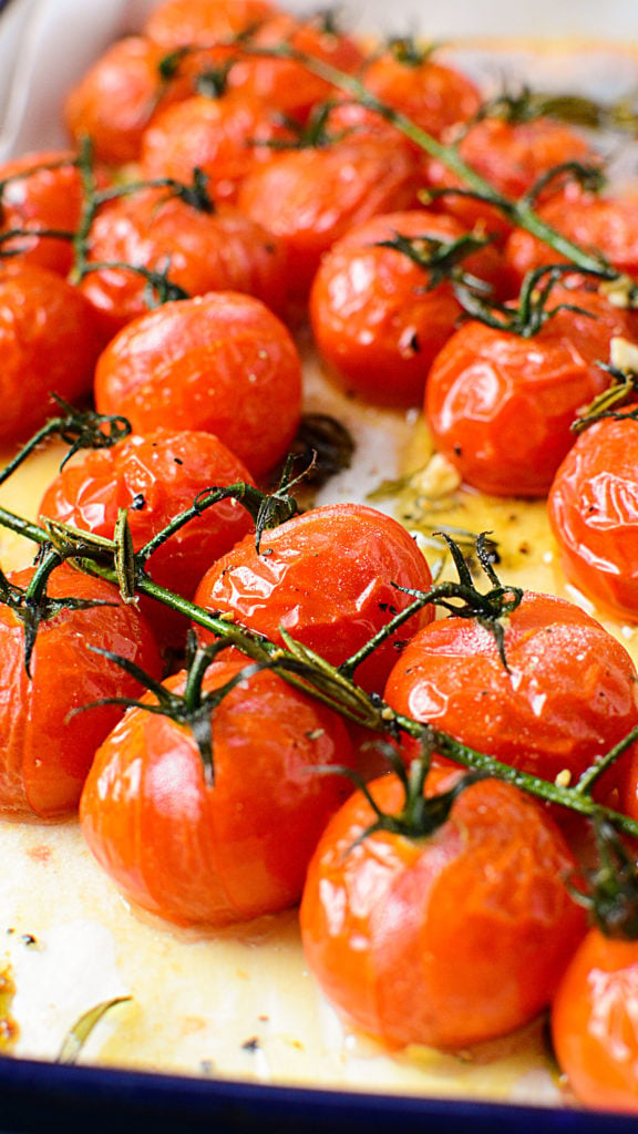 Roasted red tomatoes on a vine sitting in a white baking dish with oil and seasonings around it