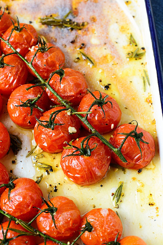 Roasted red tomatoes on a vine sitting in a white baking dish with oil and seasonings around it