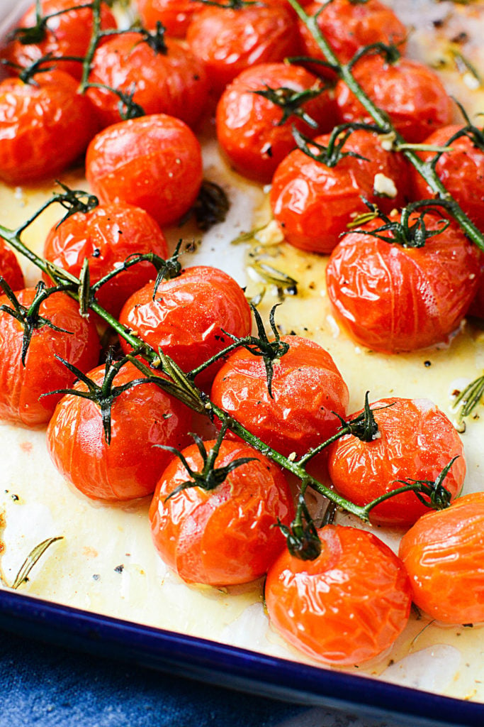 Roasted red tomatoes on a vine sitting in a white baking dish with oil and seasonings around it