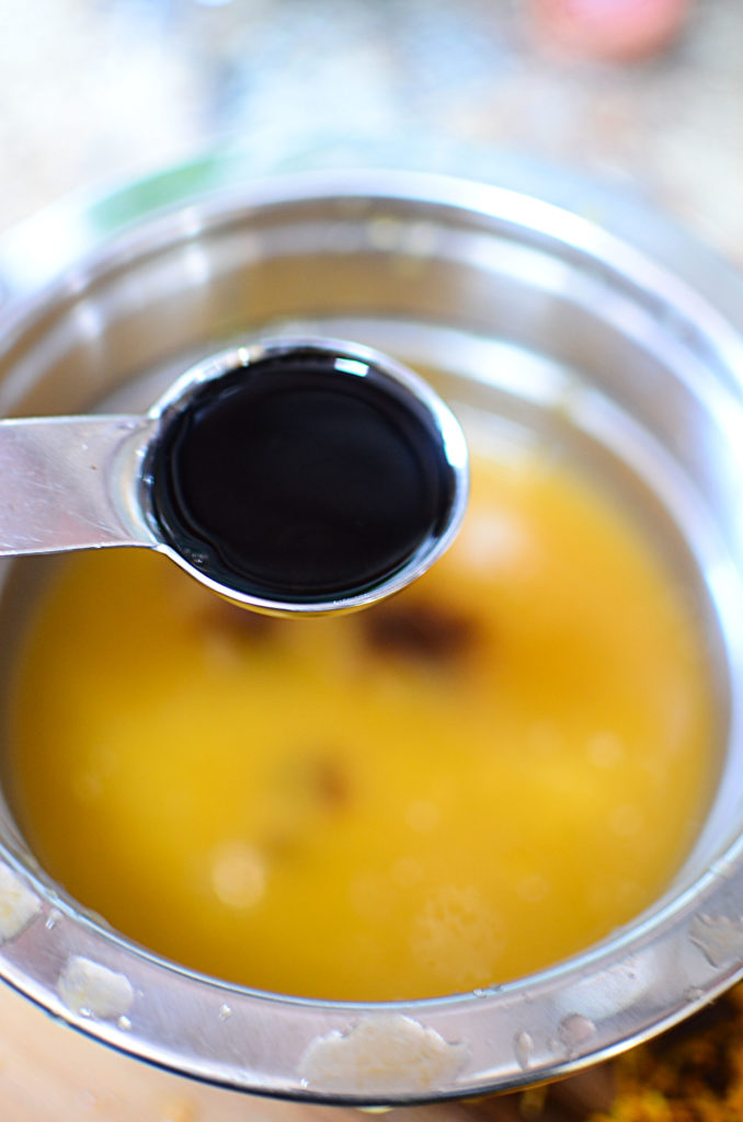 A close up image of a measuring spoon (1 teaspoon) full of honey. Background is a bowl of some orange juice.