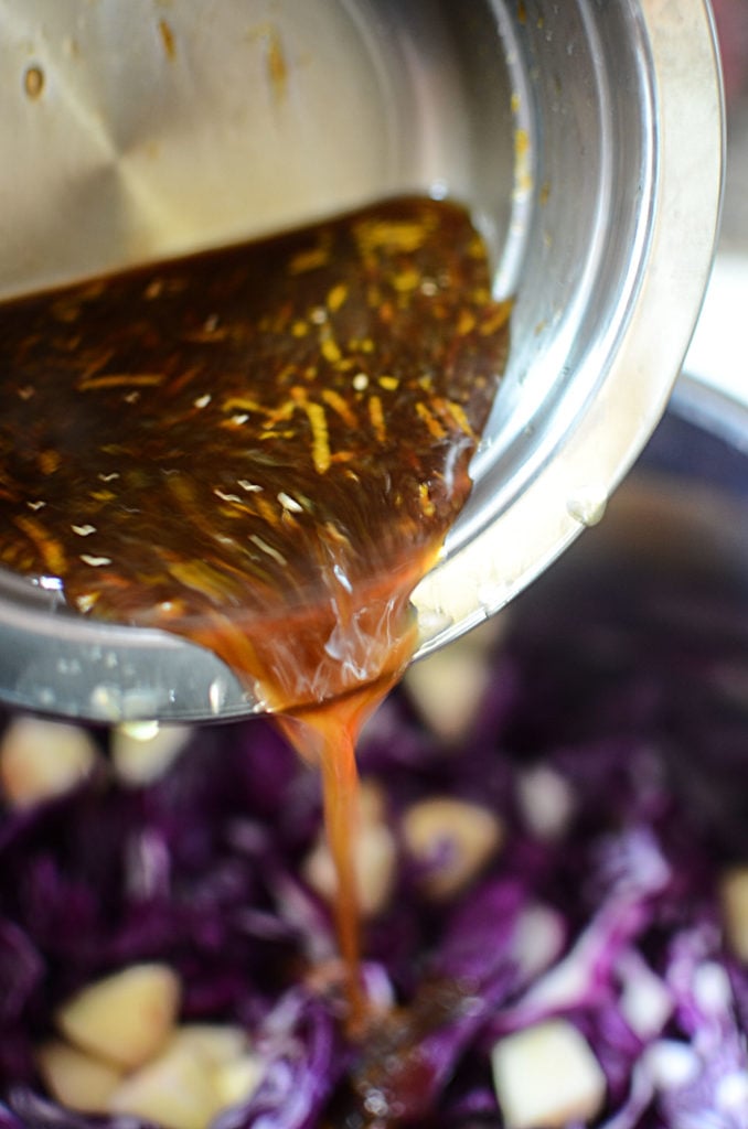 Orange juice mixture with honey and some orange zest being poured from a small stainless bowl to a bowl of chopped red cabbage and diced apples.