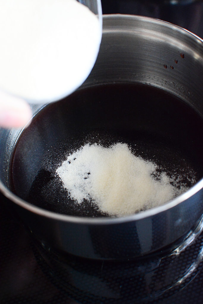A pot of elderberry syrup with warm water and a small bow of gelatin powder is poured onto the pot 