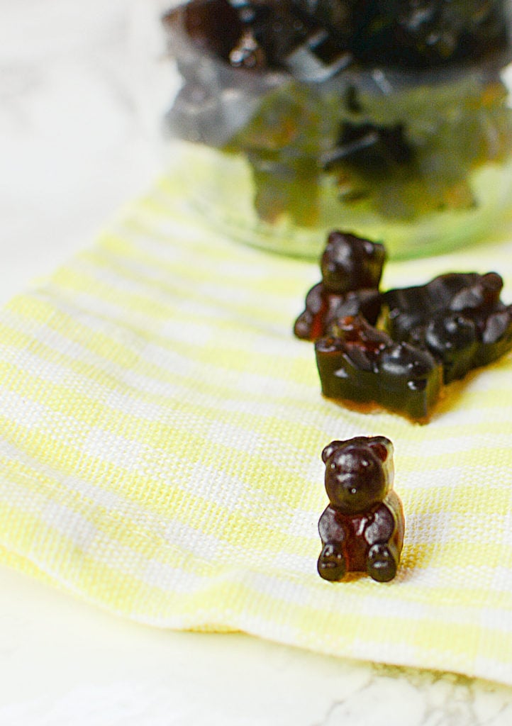 An image of elderberry syrup gummies shaped like little brown bears laid on a plaid yellow and white piece of cloth. Background is a bowlful of elderberry gummies