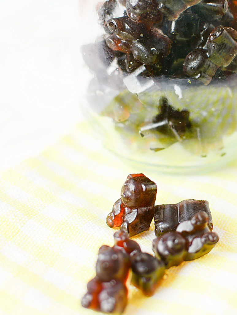 An image of elderberry syrup gummies shaped like little brown bears laid on a plaid yellow and white piece of cloth. Background is a bowlful of elderberry gummies