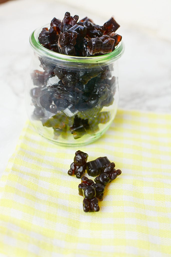 An image of elderberry syrup gummies laid on a plaid yellow and white piece of cloth. Background is a bowlful of elderberry gummies