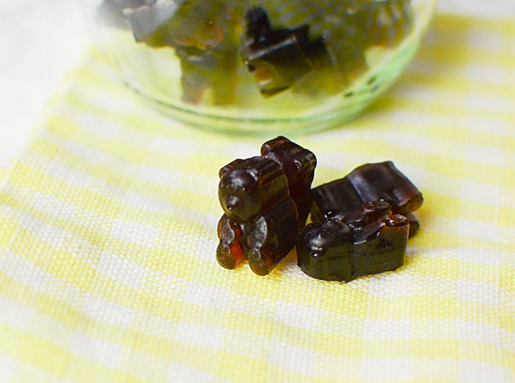 An image of elderberry syrup gummies shaped like little brown bears laid on a plaid yellow and white piece of cloth. Background is a bowlful of elderberry gummies