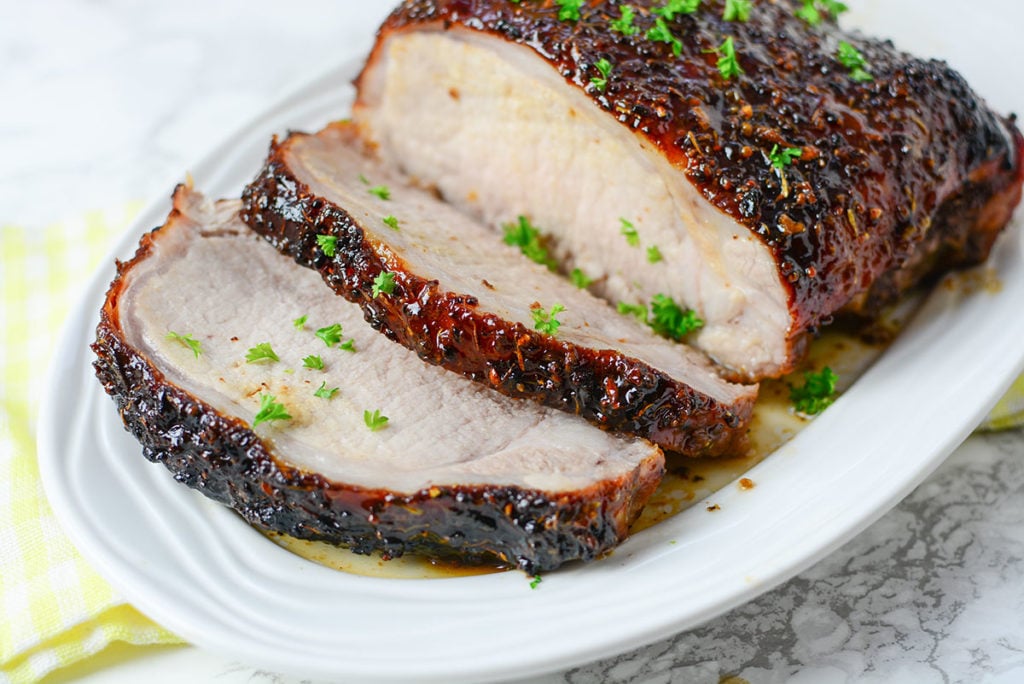 An image of honey mustard glazed piece of pork garnished with some chopped parsley. Meat if sitting on a white platter. 