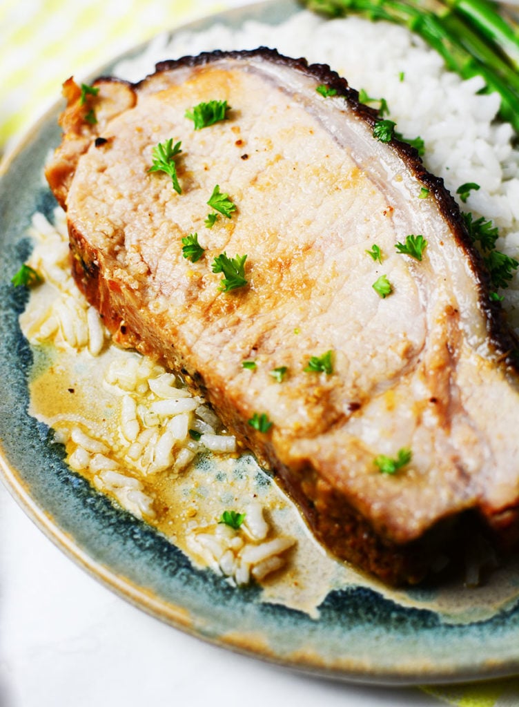 An up close image of a piece of honey mustard glazed port on top of steamed rice with some asparagus on the side, served on a blue plate.