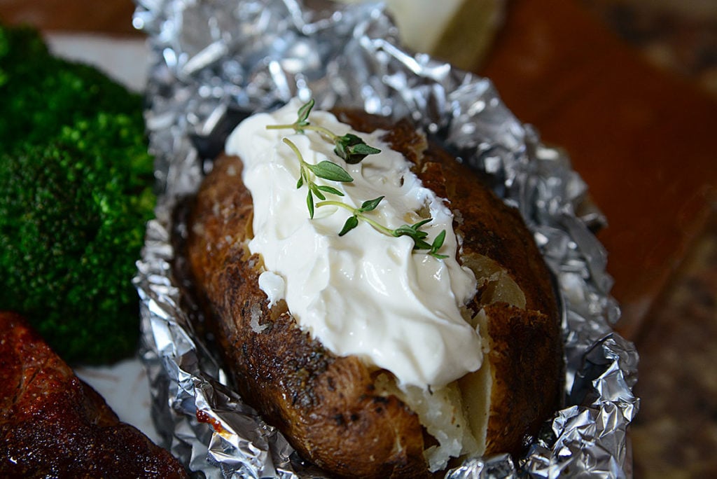 A close up image of crock pot baked potato sitting on foil and topped with some mayo garnished with some fresh herbs. Beside it is some broccoli and some piece of meat.