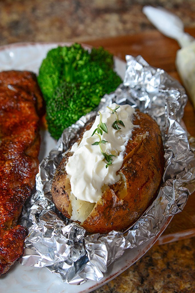 An image of crock pot baked potato sitting on foil and topped with some mayo garnished with some fresh herbs. Beside it is some broccoli and some piece of meat.