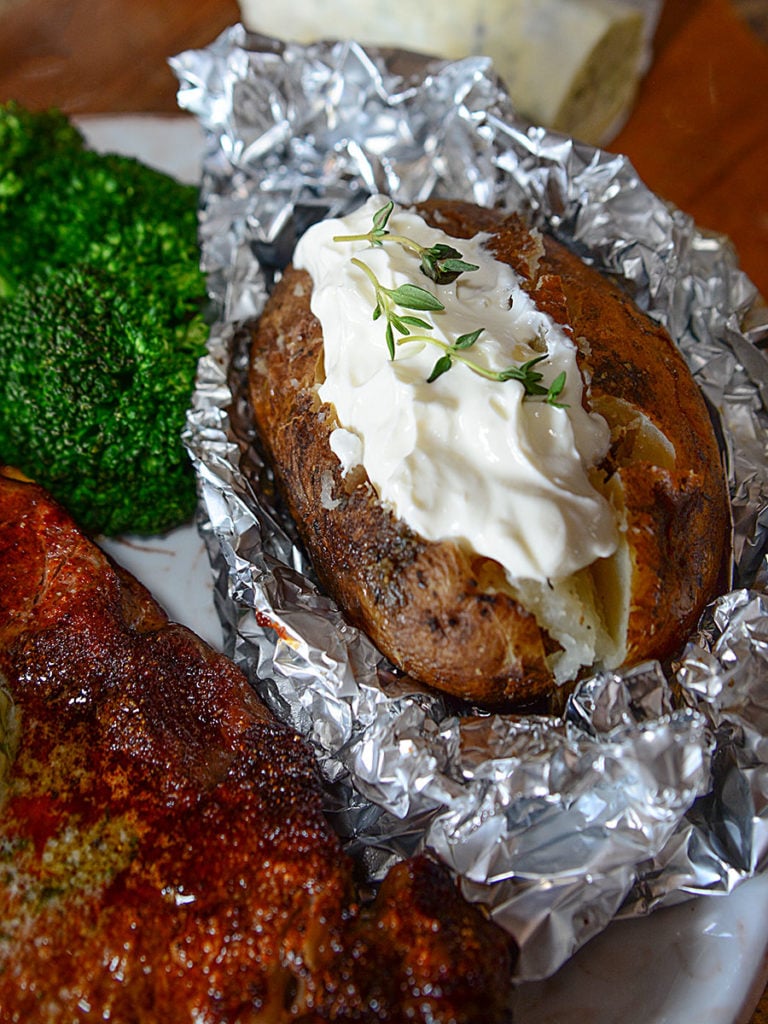 A close up image of crock pot baked potato sitting on foil and topped with some mayo garnished with some fresh herbs. Beside it is some broccoli and some piece of meat.