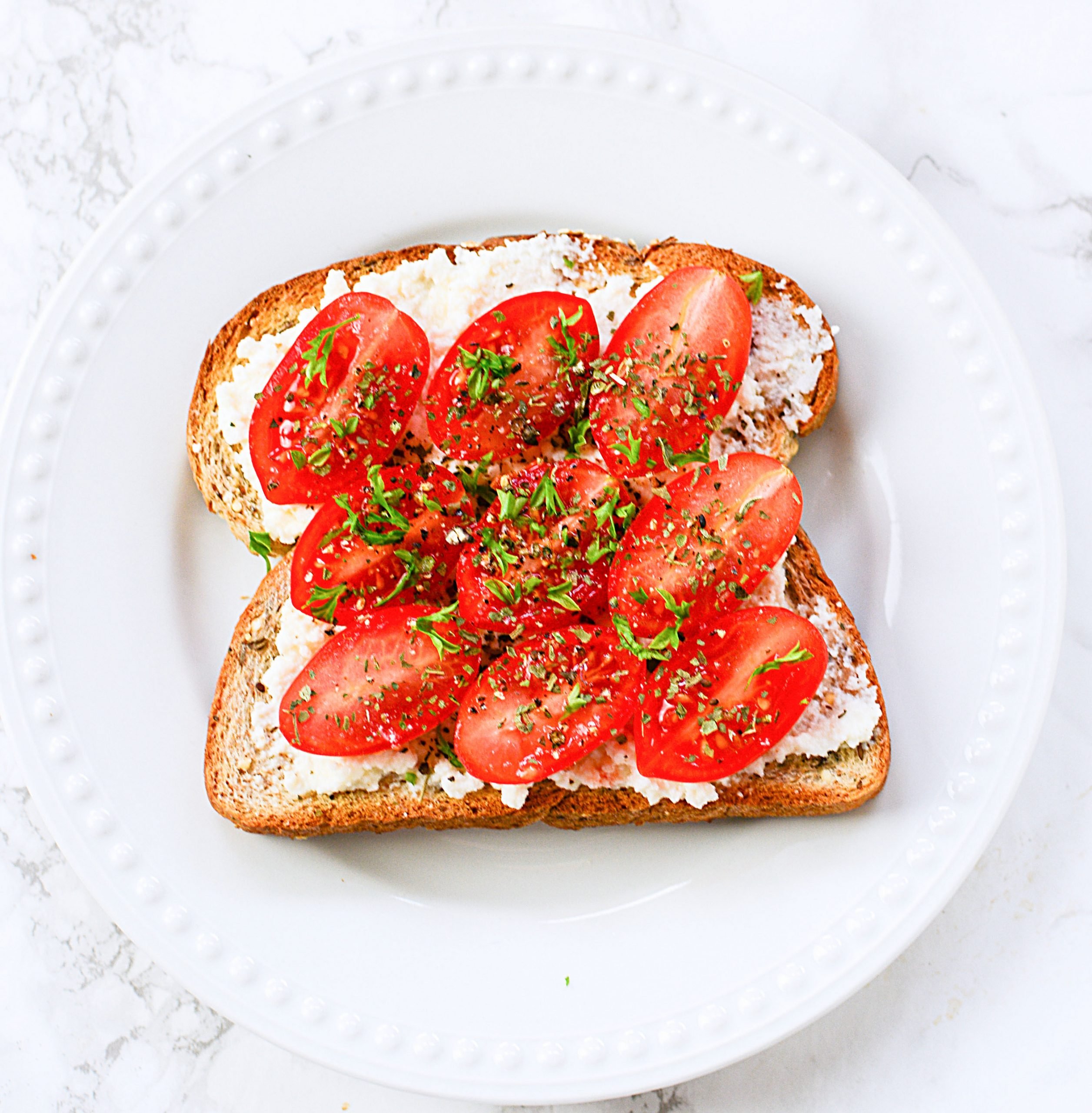 Toast on a white plate with ricotta cheese and fresh cherry tomatoes.