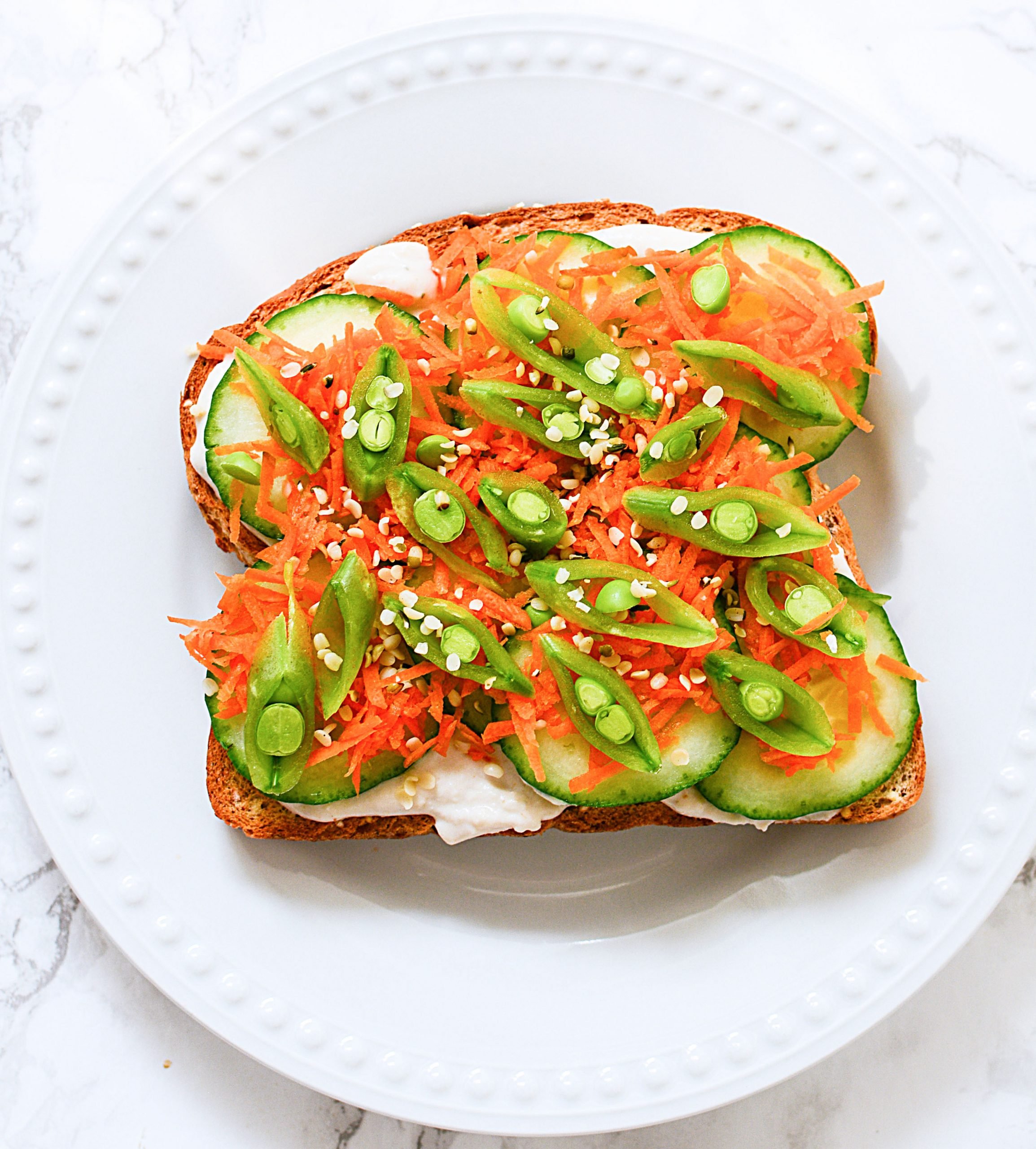 Toast with hummus, grated carrot, cucumber and sliced pea pods on top.