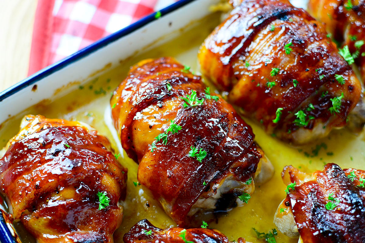 Three bacon wrapped chicken thighs on a white enamel cooking tray with blue trim.