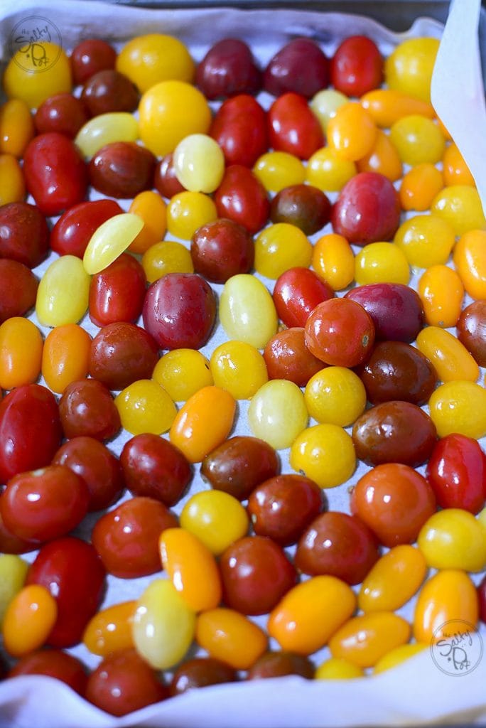 A top-down, close-up view of a baking sheet lined with white parchment paper, densely packed with raw, whole cherry and grape tomatoes in a variety of colors, including vibrant red, bright yellow, and deep orange.