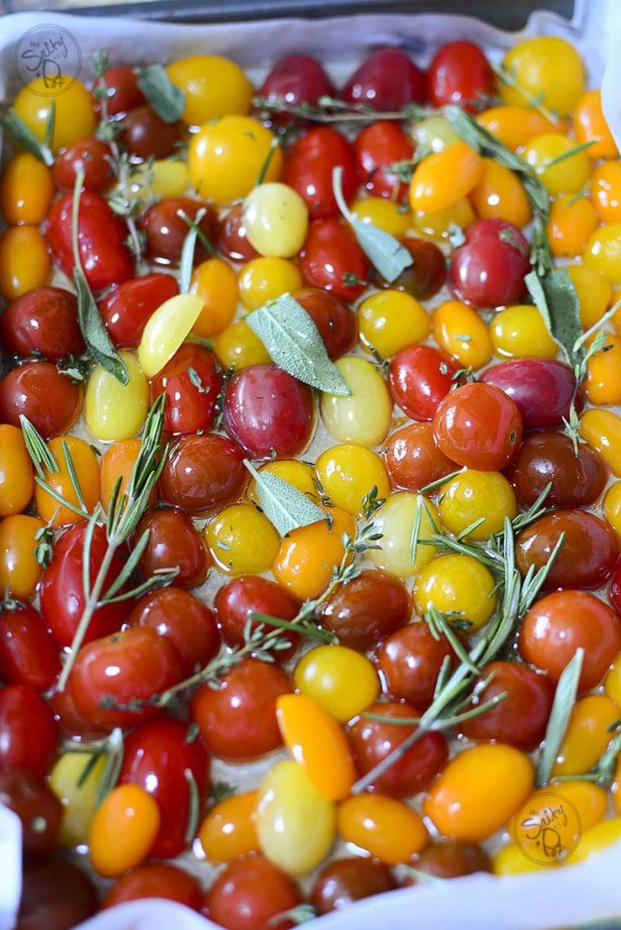 The same tray of multi-colored raw cherry tomatoes now drizzled with olive oil and topped with fresh green herbs, including sprigs of rosemary, thyme, and whole sage leaves.
