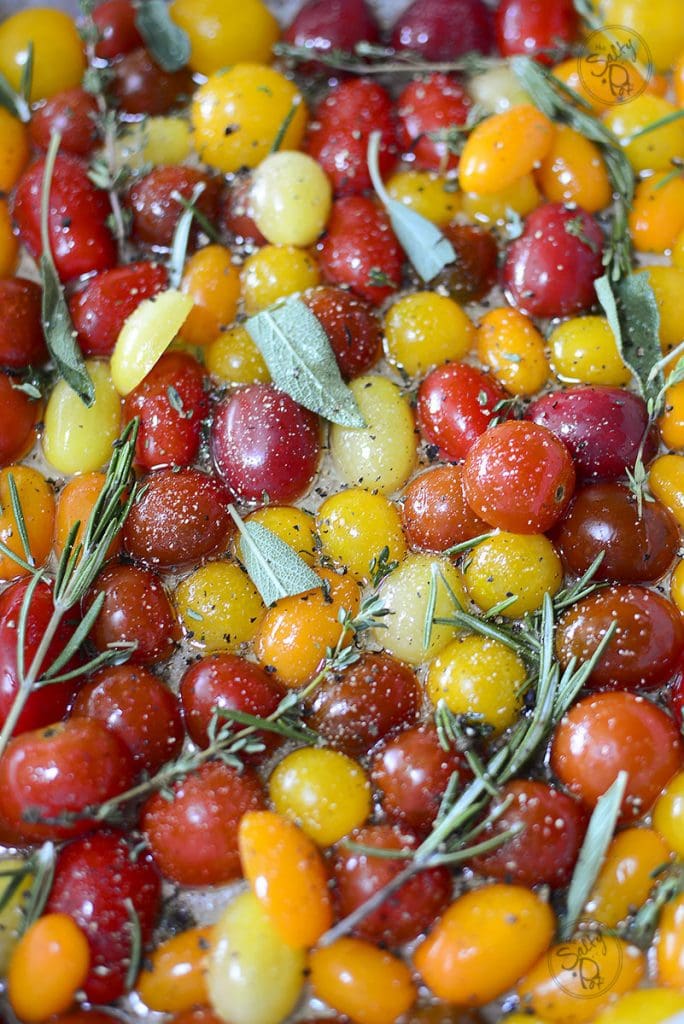 A close-up of the raw tomatoes and fresh herbs on the parchment-lined tray, now seasoned with a generous sprinkle of coarse salt and cracked black pepper.