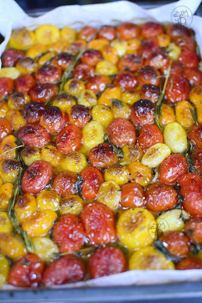 A top-down view of the roasted tomatoes after baking; the skins are blistered and slightly charred, resting in a shallow pool of infused oil and juices along with the wilted, darkened herbs.
