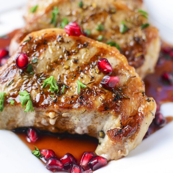 Grilled porkchops sit on a white plate with pomegranate sauce on them.