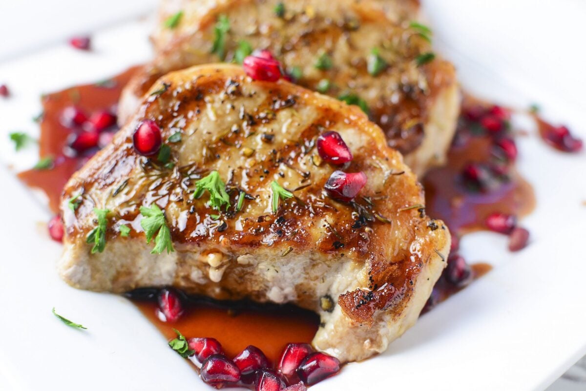 Grilled porkchops sit on a white plate with pomegranate sauce on them.