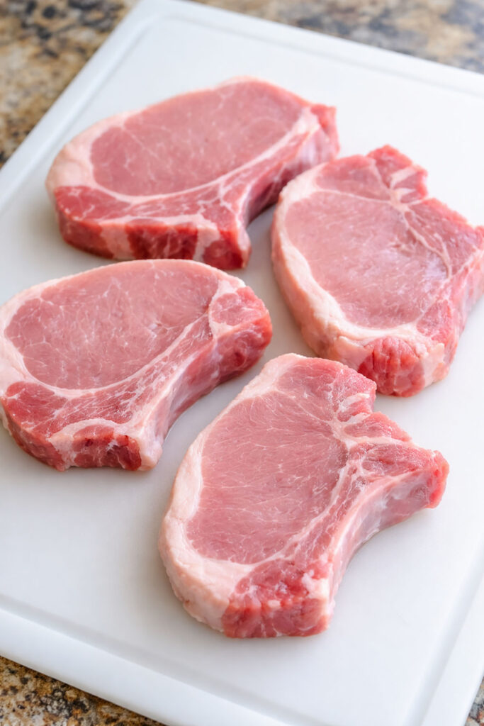 4 raw pork chops on a plastic cutting board.
