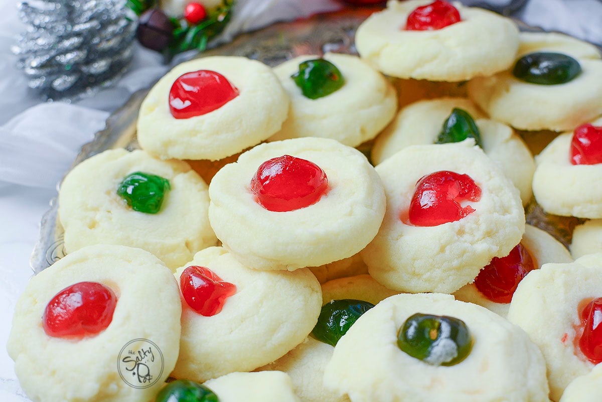 Red and Green glace cherries on whipped shortbread cookies with a silver pinecone in the background.