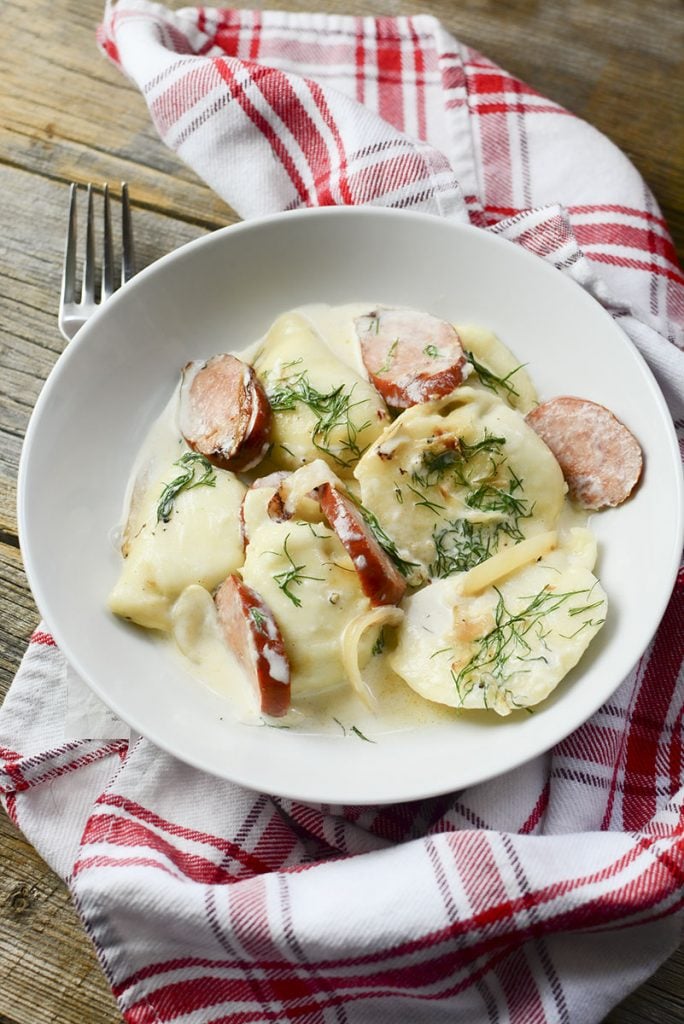 A red checked napkin sits under a bowl food of pierogies and dill in cream sauce.