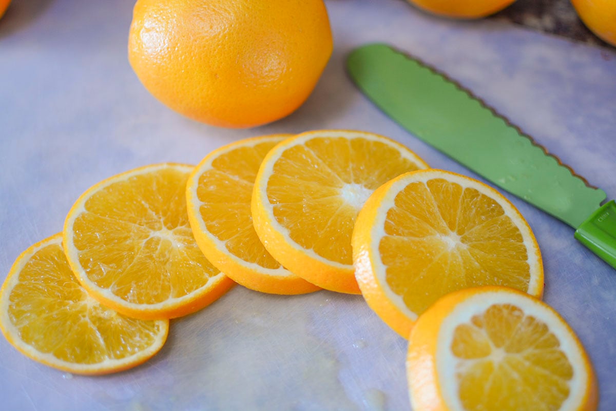 6 slices of oranges sitting next to a green serrated knife. A large whole orange sits in the background. 