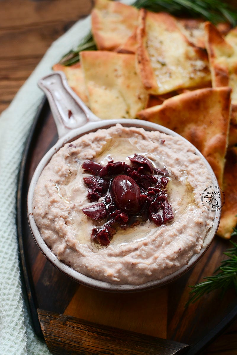 Hummus in a bowl with a handle. Black olives in the center of the hummus and baked tortilla chips in the background that are super tasty!