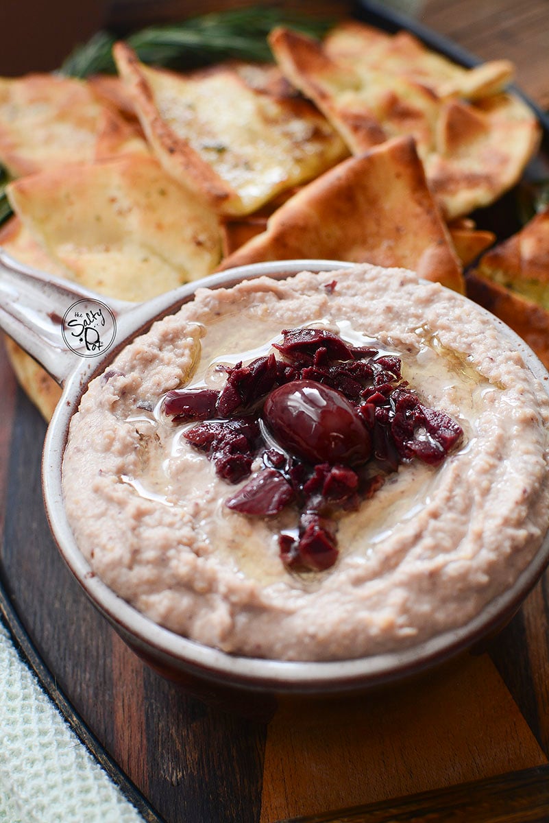 Hummus in a round crock dish with a handle, pita chips in the background.