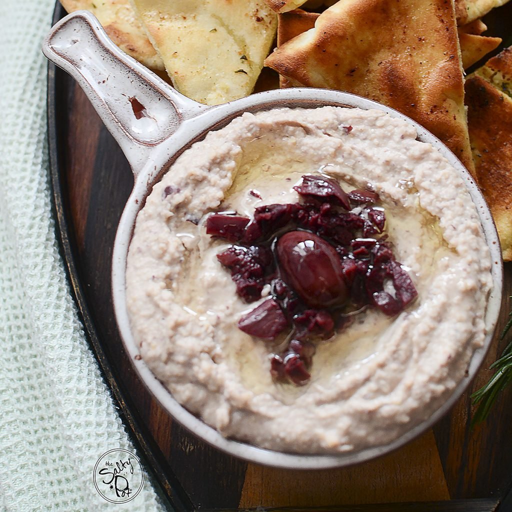 Black olive hummus in a dish with pita chips beside it.