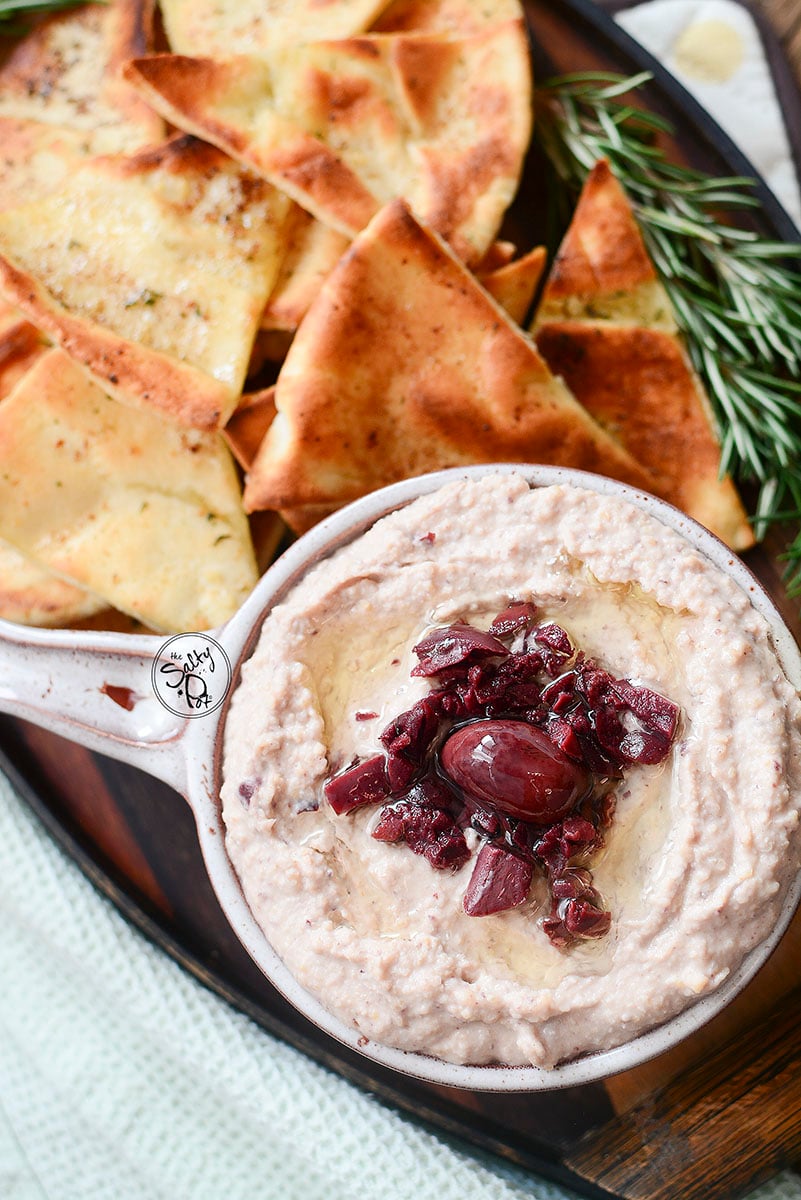 Hummus with pita chips above it, rosemary sprigs to the right, all on a wooden tray.