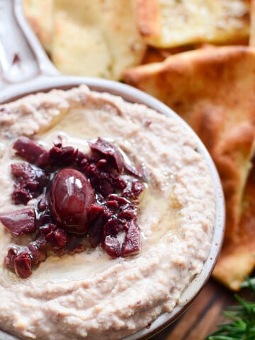 A horizontal image of the hummus in a bowl. Homemade pita chips are beside it with a sprig of fresh rosemary on the side.