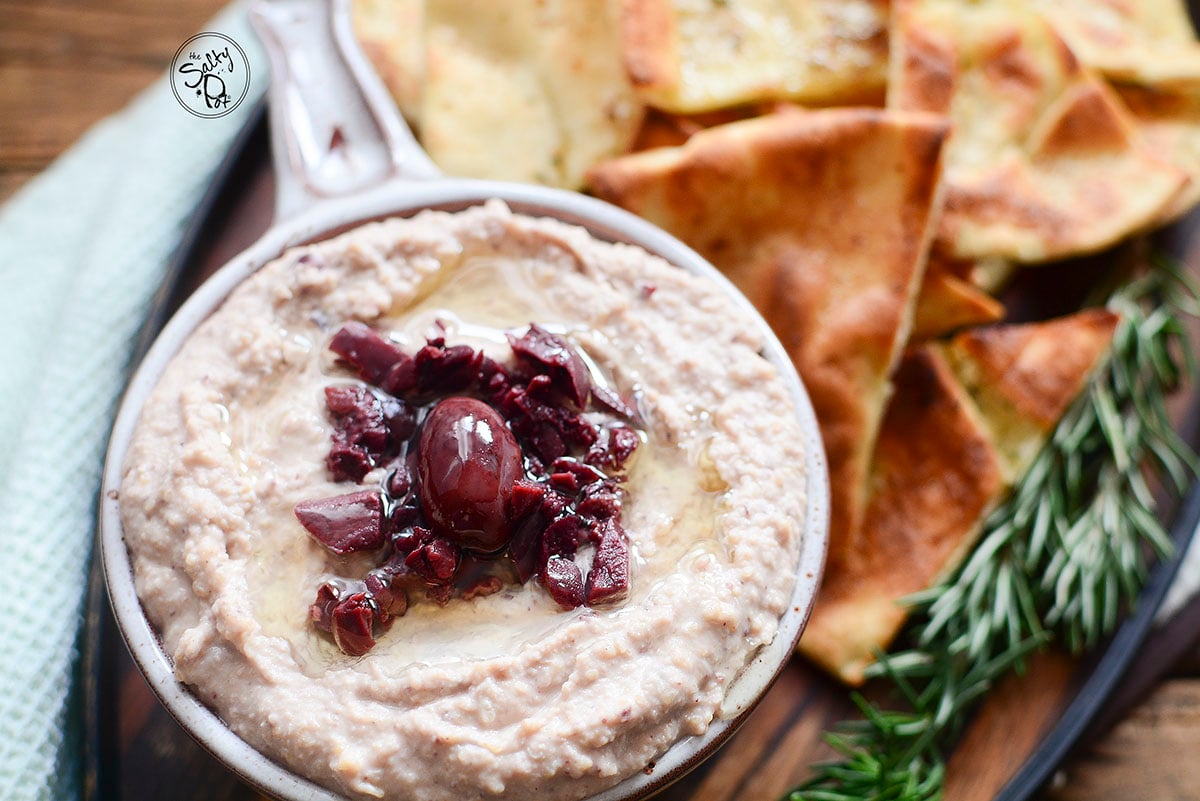 A horizontal image of the hummus in a bowl. Homemade pita chips are beside it with a sprig of fresh rosemary on the side.