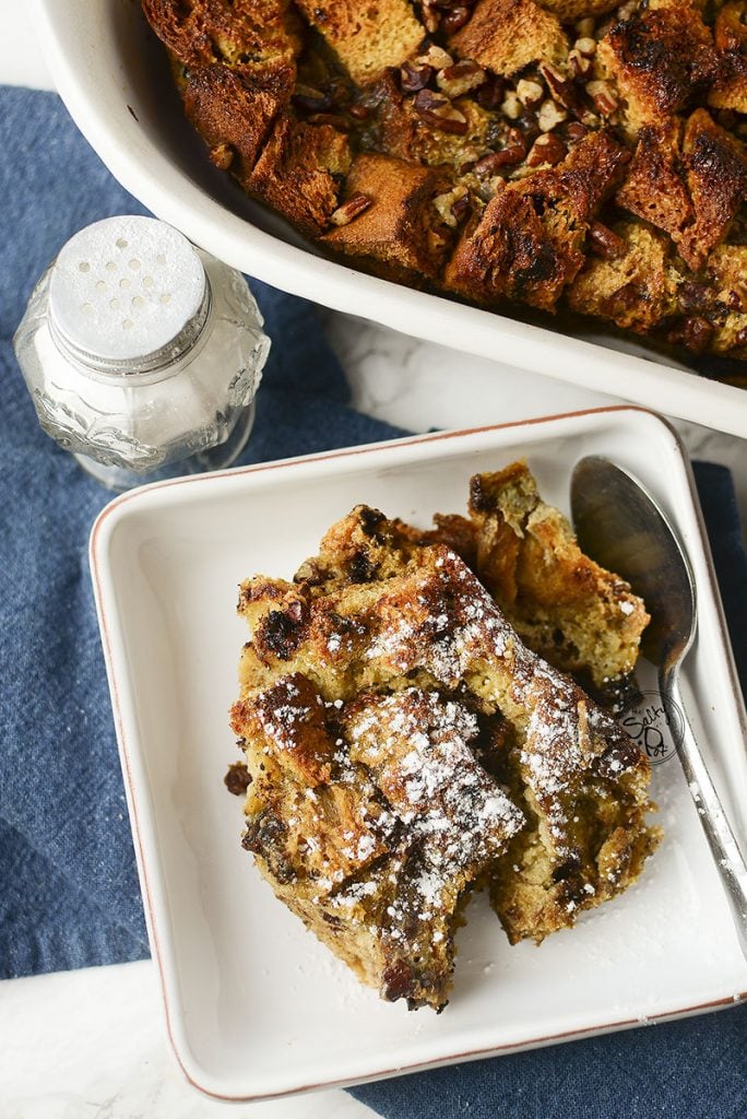 A piece of bread pudding in a white dish with icing sugar sprinkled over the top, with a spoon on the side, ready for eating!