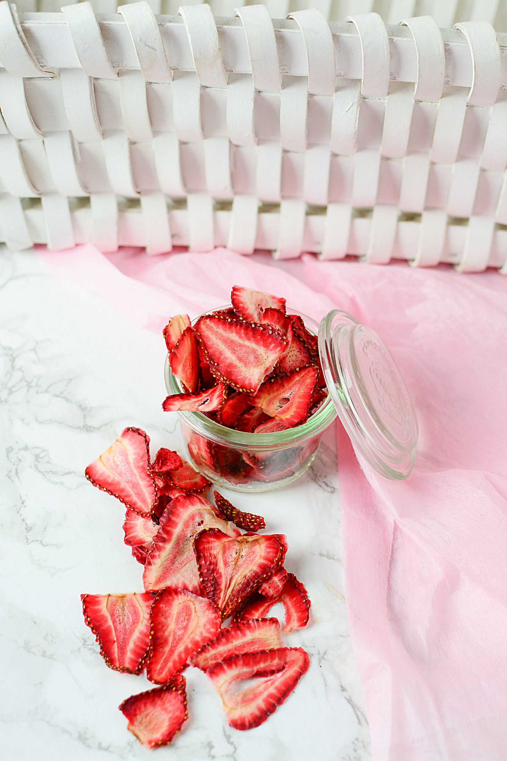 A pretty glass jar filled with overflowing dried strawberries resting close to a pink napkin. In front of the glass jar is the overflow of sliced, dried strawberries.