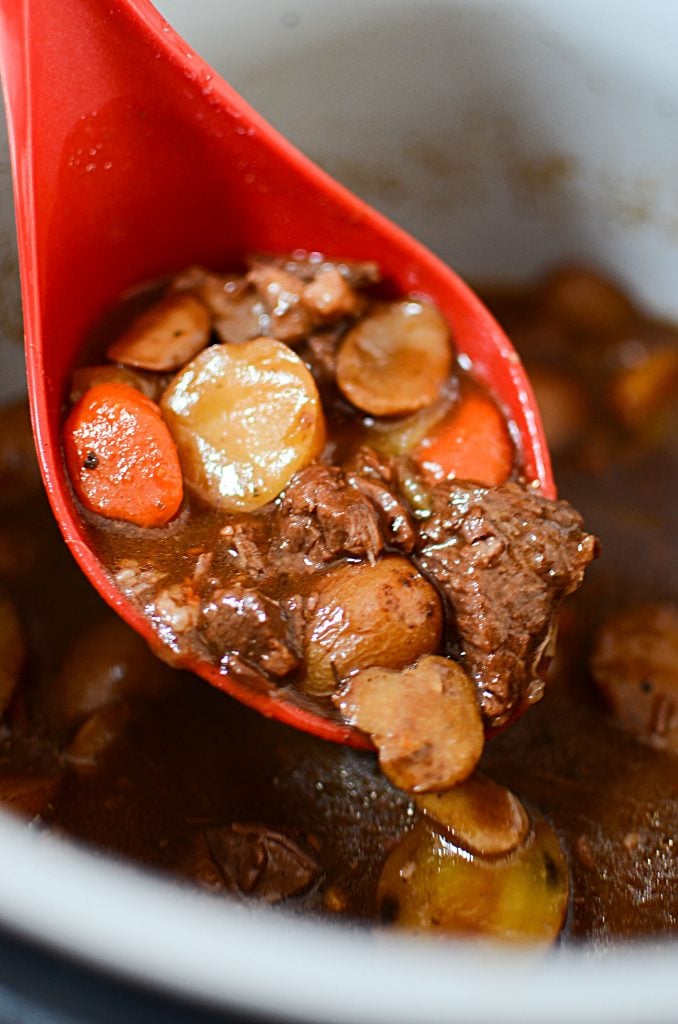 A scoop of beef stew with carrots and potatoes sitting in a red ladle.