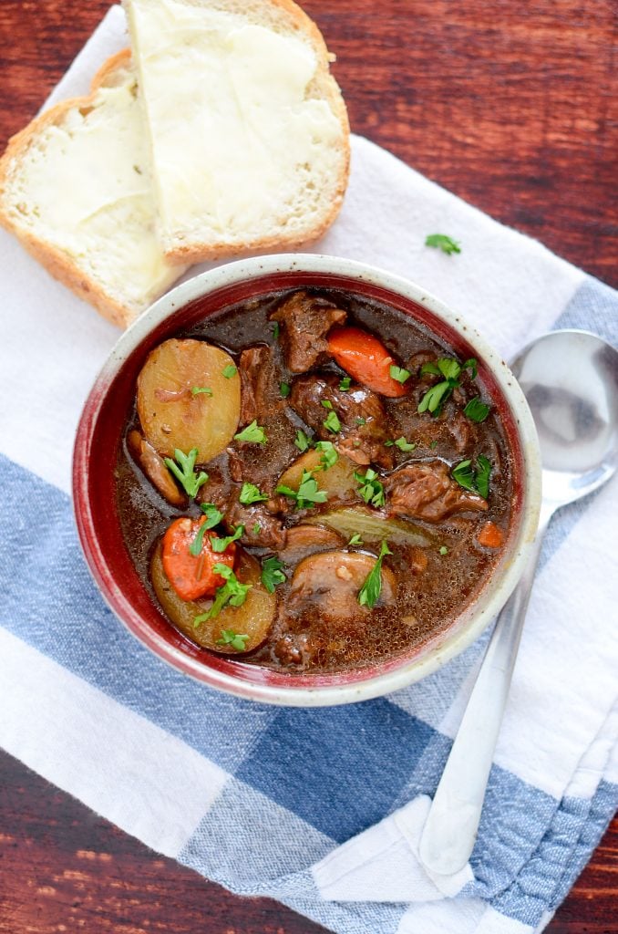 This delicious stew in a pottery bowl has a spoon sitting next to it, sitting on a white and blue napkin. 