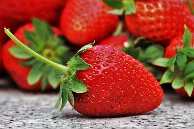 A closeup strawberry with a long stem, sitting in front of a pile of other strawberries.