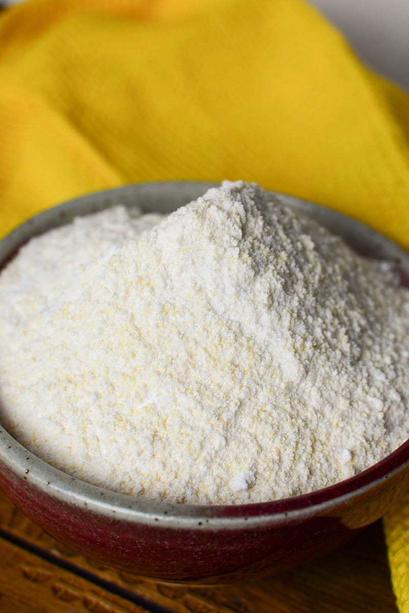 The cornbread mix is in a pottery bowl, on a wood table with a yellow napkin behind it.