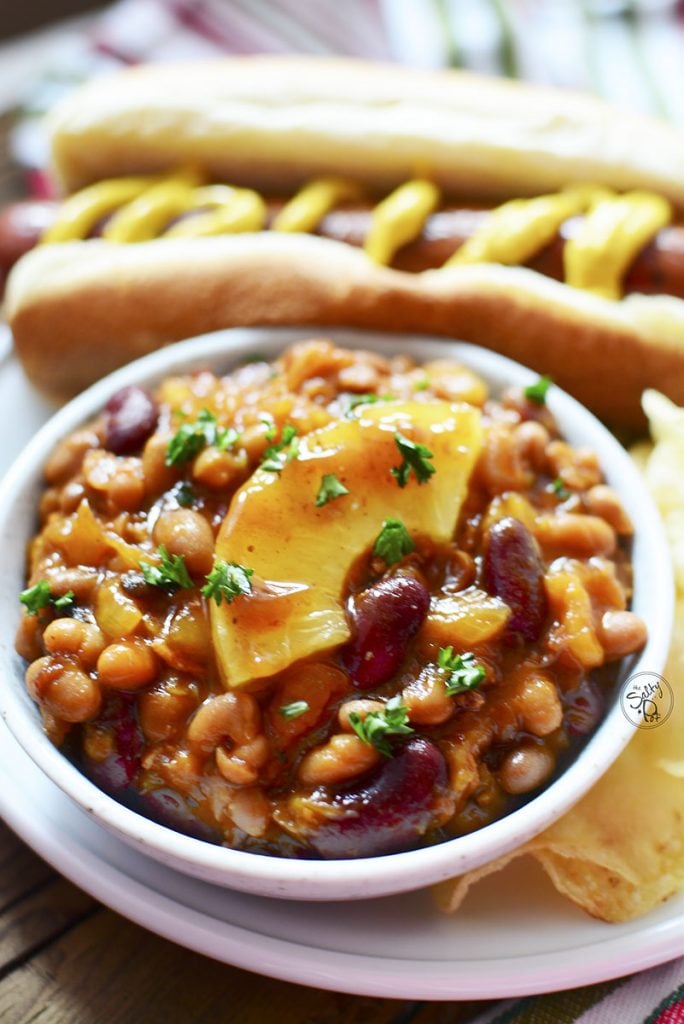 In a white bowl, a serving of the beans sit with a quarter section of pineapple sitting on top. A hot dog sits in the background.