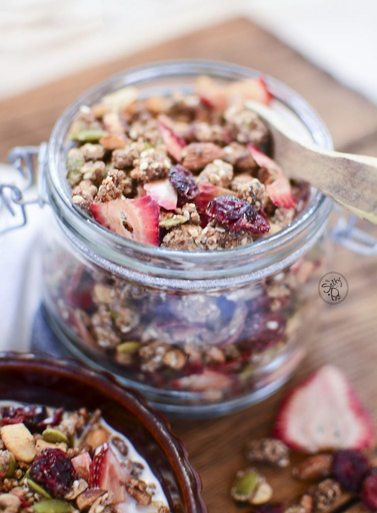 A glass container holding the granola with a small wooden spoon on top.