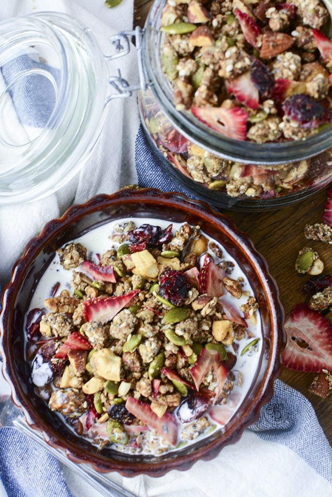 A brown bowl holding a delicious serving of the granola with milk. The container holding the bulk of the granola is off to the upper right side.