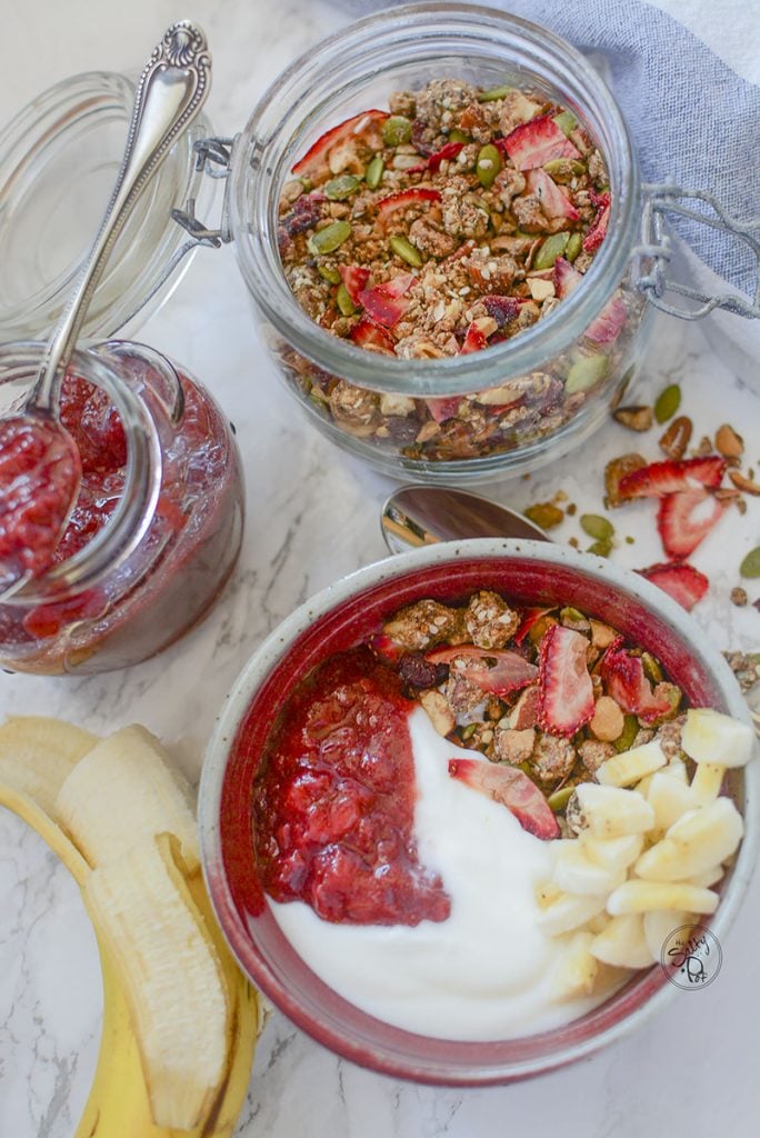 Another photo of the granola bowl breakfast spread. The granola bowl is on the bottom with a half peeled banana on the left. A spoon rests on top of the strawberry compote jar and the jar of granola sits top right.