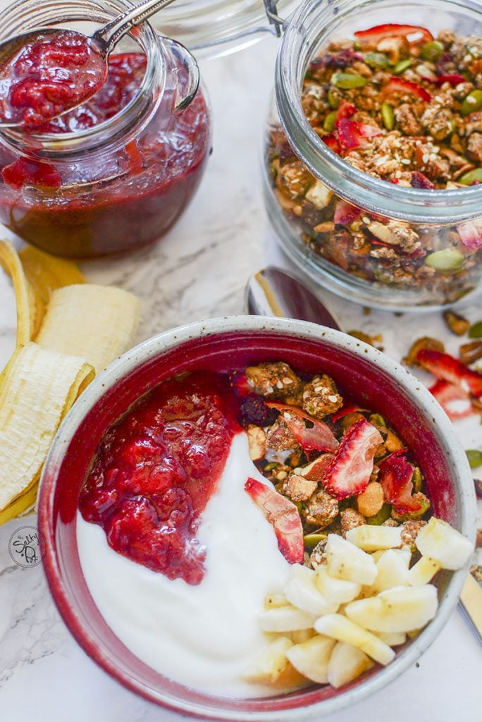 A red bowl at the bottom of the photo shows the granola bowl with strawberry compote, yogurt, and bananas on top. At the top of the photo is a jar of the strawberry jam on the left, and a jar of the granola on the right. 